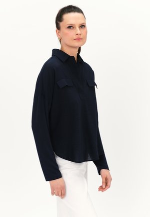 Marineblaues Langarmhemd mit Kragen, zwei Brusttaschen und lockerer Passform, kombiniert mit weißen Hosen.