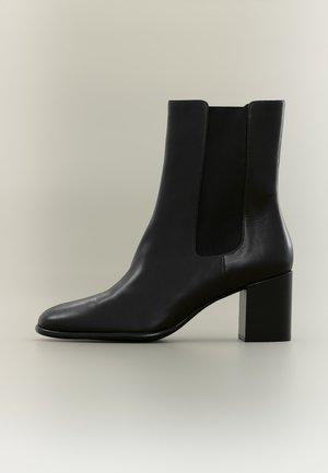 Bottine noire en cuir à bout arrondi, avec un empiècement latéral élastique et un talon bloc épais, sur fond uni.