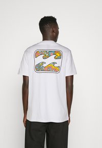 Bílý bavlněný tričko s barevným abstraktním vlnitým designem na zadní straně, s odstíny modré, růžové a žluté v obdélníkovém rámečku.