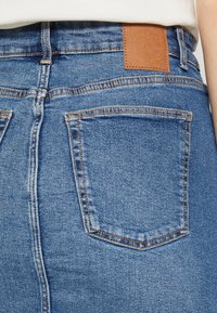 Blå denimjeans med hög midja, två bakfickor och en brun läderlapp vid midjan. Kontraststickningar finns.