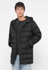 Musta untuvatakki hupulla, jossa on tikattu muotoilu, vetskari ja sivutaskut, päällä tumman värinen paita ja vaaleat farkut.