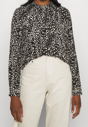 Person wearing a long-sleeve black and white leopard print blouse tucked into high-waisted off-white pants, standing against a plain background.