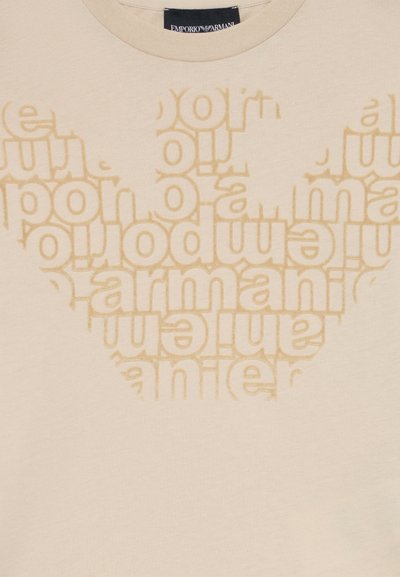 Krēmkrāsas T-kreklis ar bēšas krāsas Emporio Armani logotipu, kas veidots no atkārtota zīmola nosaukuma teksta stilizētā spārna formā.