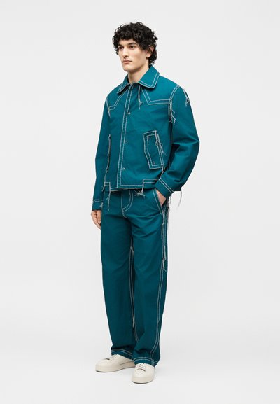 Νεαρός άνδρας στέκεται φορώντας τιρκουάζ μπουφάν και παντελόνια με αντίθετες λευκές ραφές και φθαρμένα νήματα, σε συνδυασμό με λευκά αθλητικά παπούτσια.