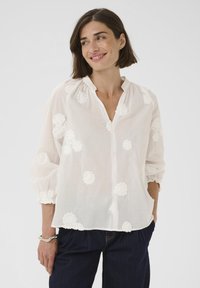 Weiße, leichte Bluse mit bestickten Blumenakzenten, einem V-Ausschnitt und langen Puffärmeln. Kombiniert mit dunkelblauen Jeanshosen.