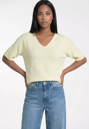 Kobieta w bladoróżowej bluzce z krótkim rękawem i dekoltem w serek oraz w wysokich niebieskich dżinsach, stojąca z rękami na biodrach na białym tle.