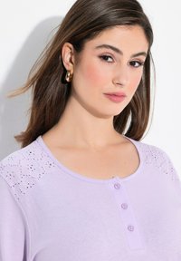 Top de color lila claro con mangas cortas que presenta detalles de encaje bordado en los hombros y una tapeta de cuatro botones en el escote. Textura suave.