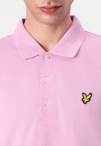 Rosa Polo-Shirt aus strukturiertem Stoff, mit einem Kragen, vier Knöpfen und einem schwarz-gelben Adler-Logo auf der linken Seite.
