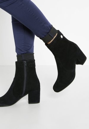 Person, die marineblaue Hose und schwarze Wildleder-Stiefeletten mit Blockabsätzen und Seitendrehverschlüssen trägt, steht auf einem weißen Boden vor einem weißen Hintergrund.