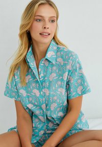Camisa de botones de manga corta en color turquesa con un estampado floral en rosa y blanco. Presenta un cuello tipo punta y está fabricada con una tela suave y ligera.
