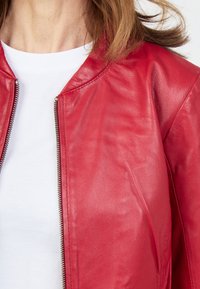 Röd läderjacka med dragkedja, rund halsringning och mjuk textur. Bärs över en vit t-shirt. Inga distinkta mönster eller accenter synliga.