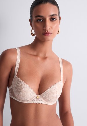 Femme aux cheveux foncés tirés en arrière portant un soutien-gorge en dentelle crème et des boucles d'oreilles créoles dorées sur un fond clair uni.