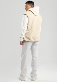 Beige Fleece-Weste über einem weißen Hoodie getragen, kombiniert mit hellgrauen Jogginghosen und weißen Sneakern, von hinten betrachtet.