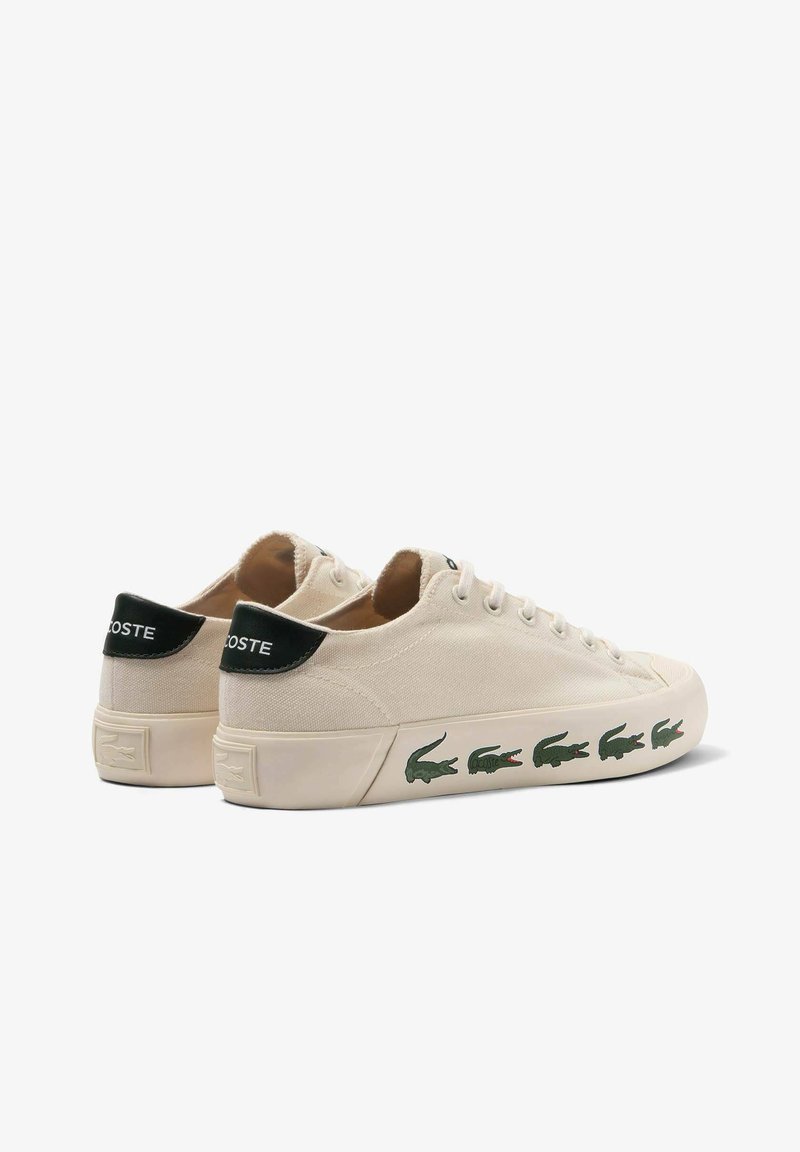 Sneaker in tela beige con forma arrotondata, accenti verdi sul tab del tallone nero e logo di coccodrillo verde ripetuto lungo la suola esterna.