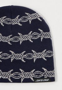 Tengerészkék kötött sapka világosszürke drótkerítéssel mintázva és egy fekete címke "Santa Cruz" felirattal, fehér háttéren.