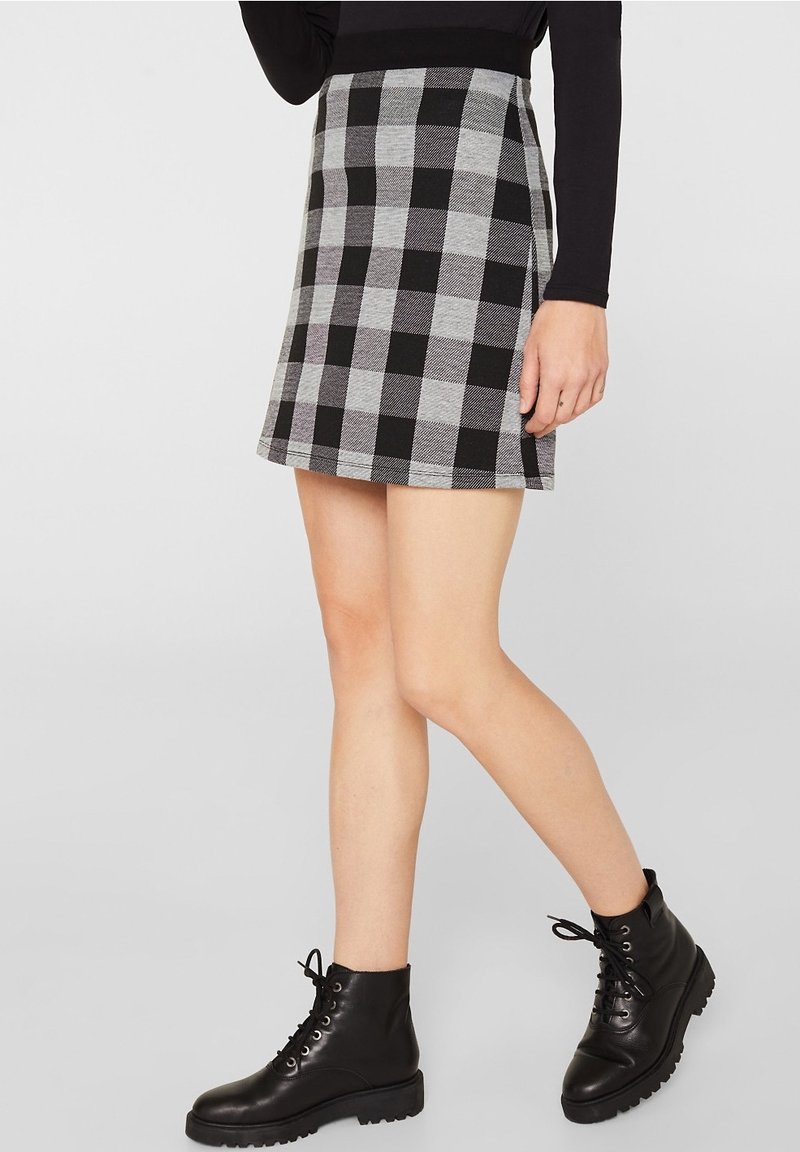 Femme portant une jupe à carreaux noire et grise, un haut noir à manches longues et des bottines à lacets noires, debout devant un fond uni.