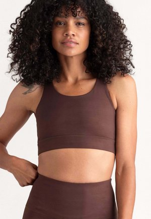 Woman with curly hair wearing a brown sports bra and high-waisted leggings, standing with one hand on her hip against a plain background.