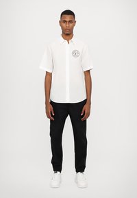 Chemise blanche à manches courtes avec un col boutonné, ornée d'un patch logo noir sur la poitrine, associée à un pantalon noir et des baskets blanches.