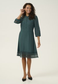 Abito teal con maniche in pizzo, dettaglio di bottoni e un bordo in pizzo all'orlo. Tessuto liscio con una gonna svasata. Indossato con tacchi neri.
