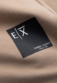Beige tyg med en slät yta, prytt med en svart rektangulär etikett med vit text: "E/X OMBRE EXIMIO SEDAN 1985" i en modern font.