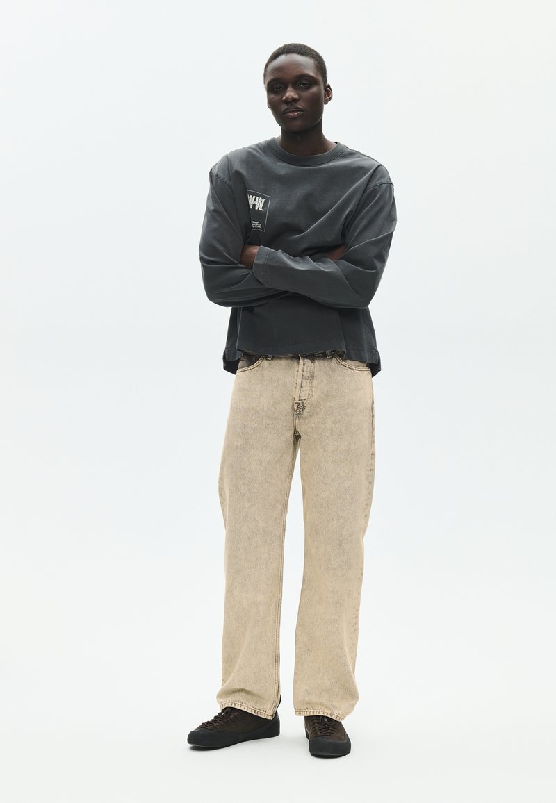 Person steht mit verschränkten Armen, trägt ein dunkelgraues langärmeliges Shirt, beige Jeans mit weitem Bein und schwarze Turnschuhe vor einem schlichten hellen Hintergrund.