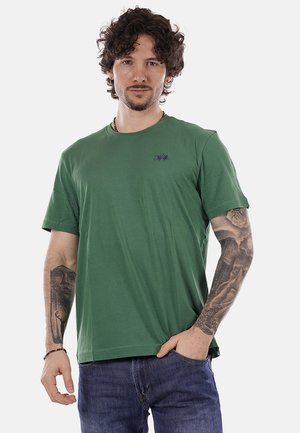 Uomo con capelli ricci che indossa una maglietta verde e jeans blu, mostra tatuaggi dettagliati su entrambi gli avambracci, con una mano in tasca.
