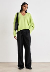 Lime greena oversized džemperis ar V veida kaklu, apvienots ar melniem platām biksēm. Modelis nes melnu somu un valkā melnas kurpes.