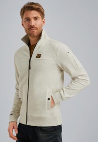 Veste beige clair zippée avec un devant texturé, un col montant, des poches latérales et un patch logo sur la poitrine. Matériau doux et design ajusté.
