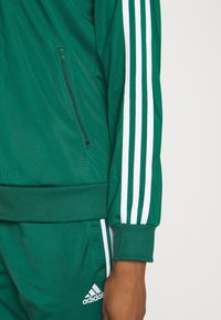 Osoba ubrana w zielony dres Adidas z białymi paskami na rękawach i nogawkach, dłoń swobodnie opuszczona wzdłuż ciała.