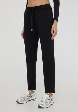 Pantaloni da tuta neri con vita regolabile, fit affusolato e texture liscia, abbinati a scarpe da ginnastica argento.