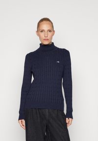 Maglione lavorato a maglia blu navy con collo alto, dotato di un design a treccia e polsini e orlo a coste. Piccolo logo sul petto.
