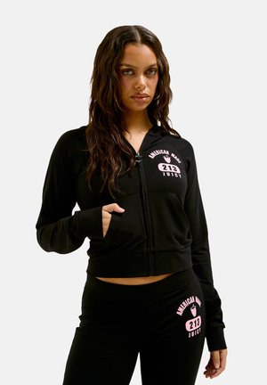 Giacca nera con zip e tasche frontali, con il logo rosa "American Made Juicy". Abbinata a pantaloni neri con lo stesso logo.