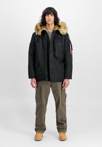 Parka neagră cu glugă căptușită cu blană, multiple buzunare și închidere cu nasturi. Purtați peste un top verde, asortat cu pantaloni cargo și botine maro.