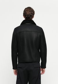Hombre con cabello corto castaño que lleva una chaqueta de borreguito negra y pantalones cargo negros, de espaldas frente a un fondo blanco liso.