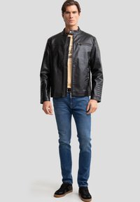 Black faux leather jacket with a mandarin collar, zip closure, and side zip pocket. Worn over a light beige shirt and blue jeans.