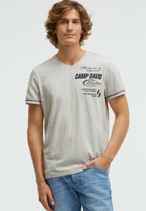 Young man with curly hair wearing a light gray short-sleeve T-shirt with black text and blue jeans, standing with one hand in pocket.