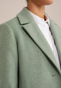 Vihreä villasekoitebleiseri, jossa on lovikaulus, jäsennellyt olkapäät ja yksirivinen nappiselkä, jota täydentää valkoinen paita alla.