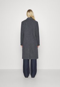 Lang, op maat gemaakte grijze jas van gestructureerde stof, met een enkele split aan de achterkant, gestanste revers en een recht silhouet over donkere wijde broekspijpen.