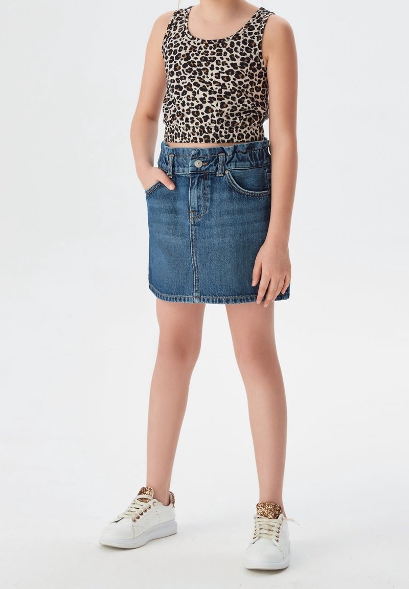 Fille portant un débardeur imprimé léopard, une jupe en jean bleue et des baskets blanches avec des touches léopard, debout avec une main dans la poche.