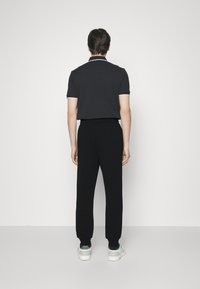 Camisa polo negra con ribetes blancos en el collar, combinada con pantalones negros ajustados. El modelo lleva zapatillas blancas. Vista posterior sobre un fondo claro.