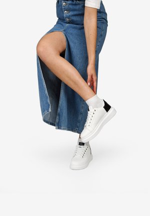 Sneaker bianche con accenti neri, materiale sintetico testurizzato, design con lacci, abbinate a una gonna di jeans con spacco laterale.