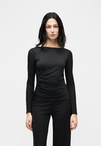 Γυναίκα με σκούρα ίσια μαλλιά, ντυμένη με εφαρμοστό μαύρο μακρυμάνικο τοπ με ρυτιδωμένες πλευρές και μαύρο παντελόνι, στεκόμενη μπροστά σε απλό λευκό φόντο.