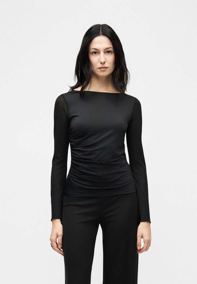 Γυναίκα με σκούρα ίσια μαλλιά, ντυμένη με εφαρμοστό μαύρο μακρυμάνικο τοπ με ρυτιδωμένες πλευρές και μαύρο παντελόνι, στεκόμενη μπροστά σε απλό λευκό φόντο.