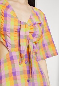 Camisa xadrez colorida em laranja, rosa, verde e roxo, com decote em V, mangas bufantes, detalhe de laço no busto e botões roxos.