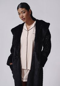Sort badekåbe i kunstpels med tekstureret design, stor krave og to sidelommer. Båret over et lyst beige sæt pyjamas med en lomme.