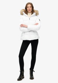 Chaqueta de plumas blanca con capucha con borde de piel. El diseño incluye un cierre frontal con cremallera y una silueta ajustada, combinado con leggings negros y botas con cordones.