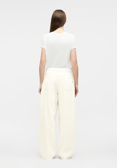 Femme aux cheveux longs et lisses, vue de dos, portant un t-shirt blanc uni à manches courtes et un pantalon blanc ample à jambes larges.