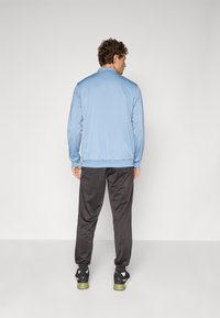 Lyseblå træningsjakke med lange ærmer og krave, parret med mørkegrå bukser og sorte sneakers, set fra ryggen. Glat stofstruktur.
