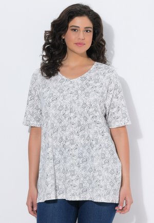 Vrouw met donker krullend haar, met hoepeloorbellen, een wit kort mouw shirt met bloemenpatroon en blauwe jeans, tegen een effen achtergrond.