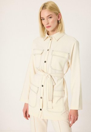 Lyshåret kvinde iført cremefarvet knapjakke med sort syning og bælte, stående mod en ensfarvet lys baggrund.